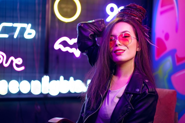 Retrato da moda de uma jovem de óculos escuros posando perto de placas de néon em uma boate