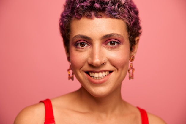 Retrato da moda de uma mulher com um corte de cabelo curto de cor roxa e um sorriso com dentes em um top vermelho em uma felicidade de fundo rosa