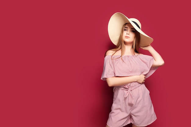 Retrato da moda e beleza de uma jovem linda em um vestido vermelho claro e um chapéu de palha em uma parede vermelha