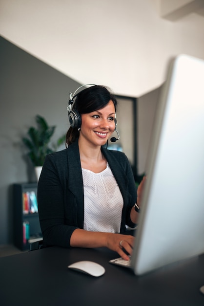 Retrato da mulher de negócios bonita com os auriculares que trabalham no computador.