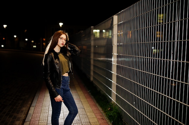 Retrato da noite do modelo menina vestir jeans e jaqueta de couro contra cerca de ferro.