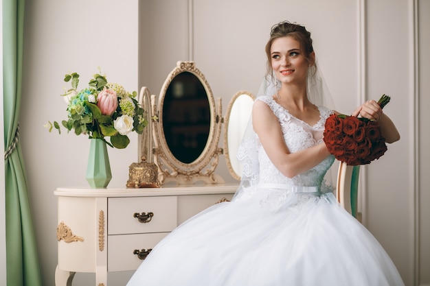 Retrato da noiva bonita no roupão de seda branco com penteado encaracolado e véu longo, sentado perto da janela no quarto, copie o espaço.