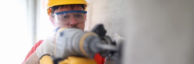 Retrato da parede concentrada da perfuração do trabalhador masculino com equipamento profissional. homem no capacete protetor amarelo. capataz de óculos. local de construção e conceito de renovação