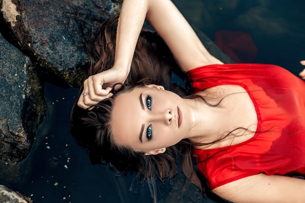 Retrato da vista de cima da sereia jovem bonita molhada deitada na rocha na água do rio ou lago tocando a cabeça vestida de vestido vermelho olhando para a câmera, cópia espaço e fundo desfocado da natureza