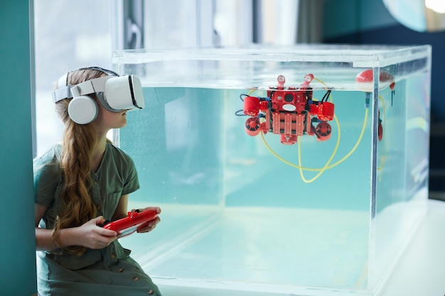 Retrato da vista lateral da menina usando fone de ouvido VR durante a operação do barco robótico no laboratório da escola, copie o espaço