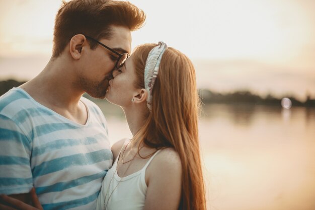 Retrato da vista lateral de um casal caucasiano encantador beijando contra o pôr do sol, perto de um lago, enquanto namorava ao ar livre.