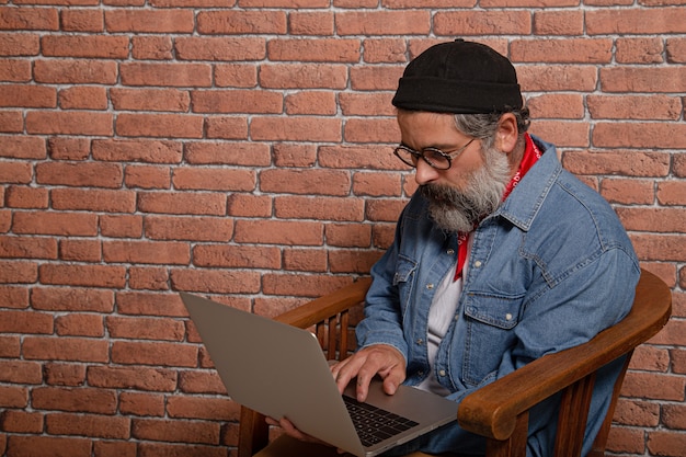 Retrato da vista lateral de um homem barbudo em camisa jeans, sentado na cadeira de madeira, trabalhando no laptop.