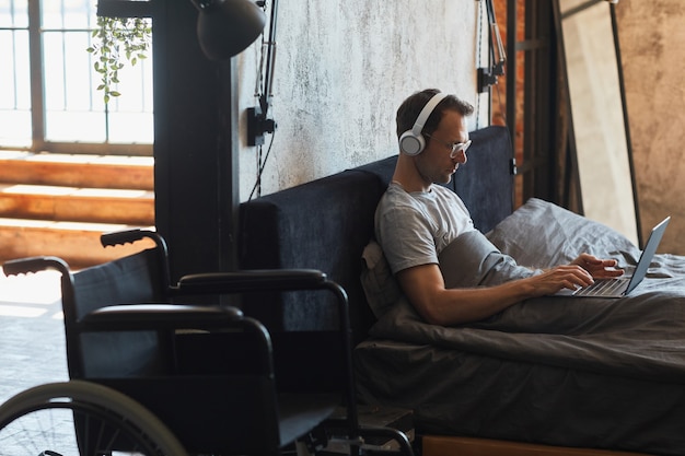 Retrato da vista lateral de um homem moderno com deficiência usando o laptop na cama, a cadeira de rodas em primeiro plano, copie o espaço