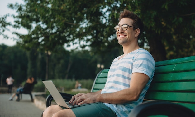 Retrato da vista lateral de um jovem freelancer masculino confiante trabalhando em seu laptop enquanto está sentado em uma praia no parque, olhando para longe, sorrindo.
