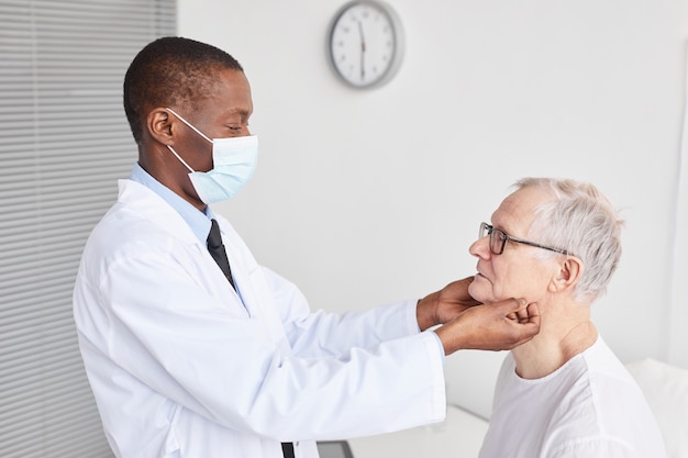 Retrato da vista lateral de um médico afro-americano examinando um homem sênior em um quarto branco de hospital, copie o espaço