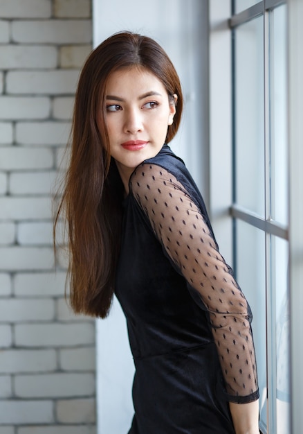 Retrato da vista lateral de uma encantadora mulher asiática, da etnia Laos, em um elegante vestido preto em pé com uma pose calma e fofa perto da janela e olhando para longe
