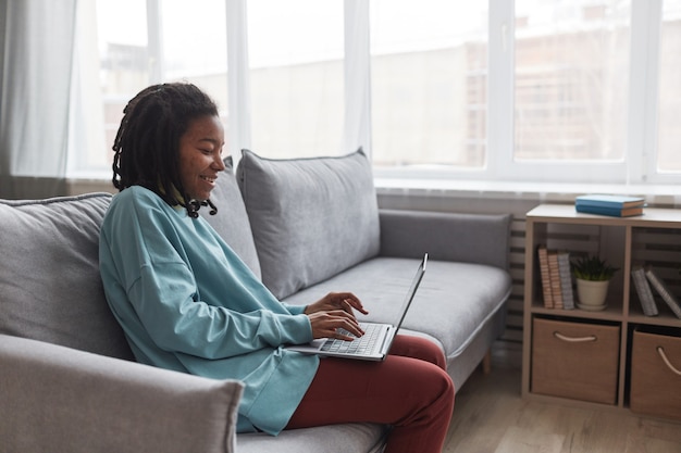 Retrato da vista lateral de uma jovem afro-americana usando o laptop e sorrindo enquanto está sentada no sofá em casa com foco nas imperfeições da pele, copie o espaço