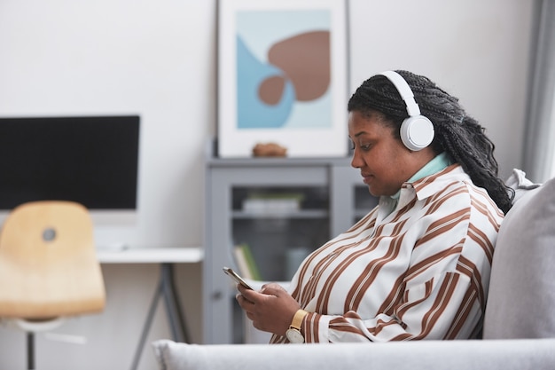 Retrato da vista lateral de uma mulher afro-americana curvilínea usando fones de ouvido e ouvindo música no smartphone enquanto está sentada no sofá no interior de uma casa mínima, copie o espaço