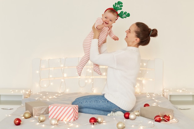 Retrato da vista lateral de uma mulher atraente com coque de cabelo, vestindo jeans e suéter branco, segurando a filha infantil nas mãos, sentada na cama, em um quarto claro com decoração de Natal.