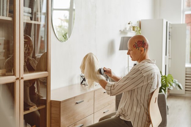Retrato da vista lateral de uma mulher careca moderna escovando a peruca enquanto está sentada perto do espelho no interior da casa, alopecia e conscientização do câncer, copie o espaço