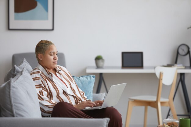 Retrato da vista lateral de uma mulher moderna de raça mista usando laptop enquanto relaxa no sofá em casa, copie o espaço