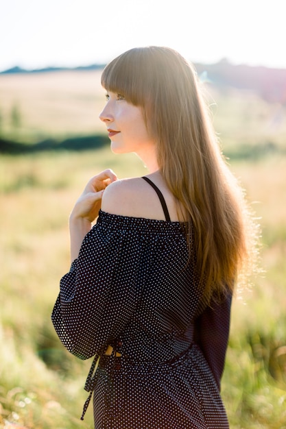 Retrato da vista lateral de uma romântica mulher caucasiana em roupas pretas, caminhando em um campo incrível de verão durante o pôr do sol quente