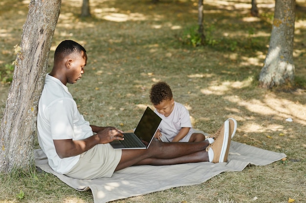 Retrato da vista lateral do pai afro-americano usando o laptop enquanto brincava com o filho bonito no parque cópia s.