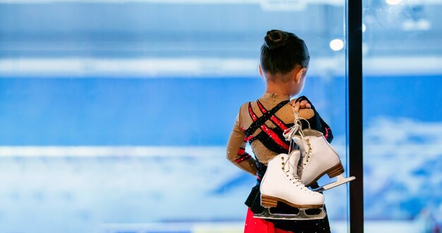 Retrato da vista traseira da menina segurando patins em pé na pista de gelo coberta