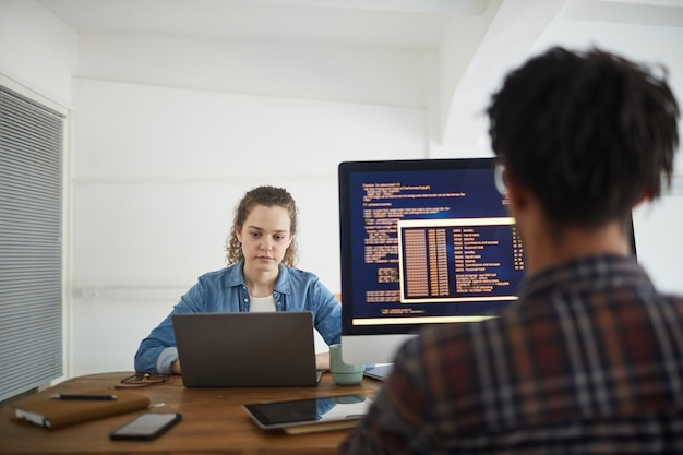 Retrato da vista traseira de um homem afro-americano escrevendo código na tela do computador enquanto trabalhava na mesa da agência de desenvolvimento de software com uma colega usando o laptop em segundo plano, copie o espaço