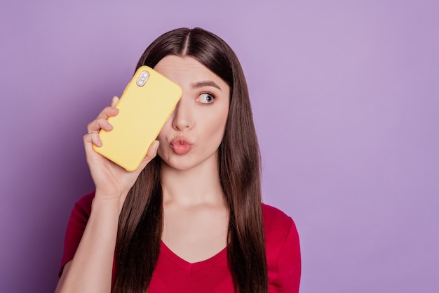 Retrato de dama adorable divertida mantenga el teléfono cerca del ojo enviar soplo aire beso mirar espacio en blanco sobre fondo púrpura