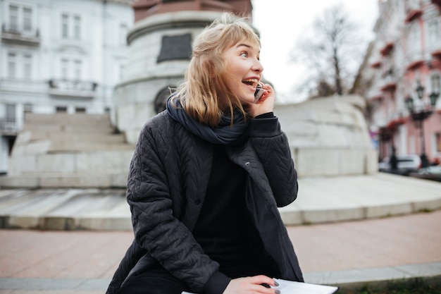 Retrato de dama alegre con cabello rubio hablando por su teléfono celular mientras mira con alegría a un lado en las calles de la ciudad