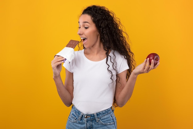 Retrato de dama emocionada sosteniendo manzana y mordiendo chocolate