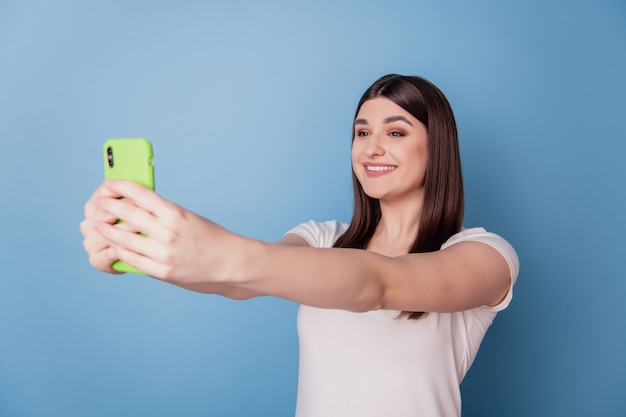 Retrato de dama feliz soñadora mantenga smartphone tomar selfie con gran sonrisa radiante sobre fondo azul.