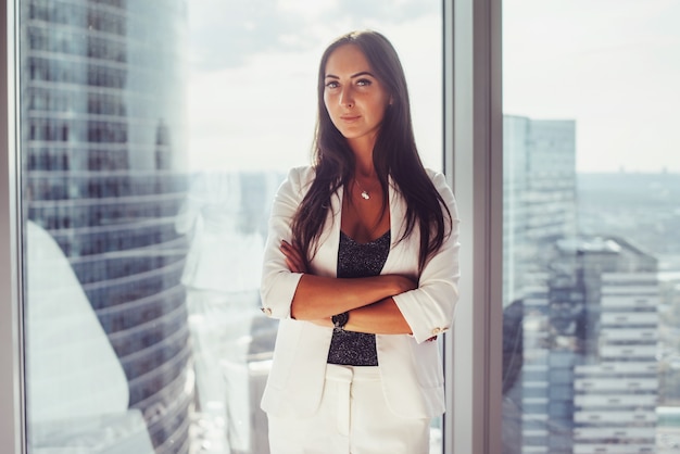 Retrato de dama de negocios elegante vistiendo traje formal blanco de pie junto a la ventana