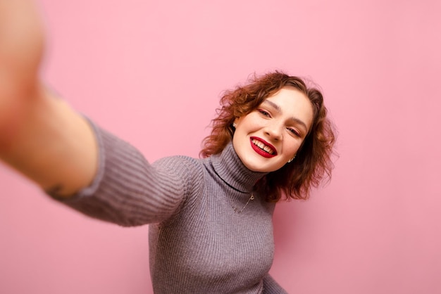 Retrato de dama rizada sonriente tomando selfie sobre fondo rosa mirando a la cámara y sonriendo