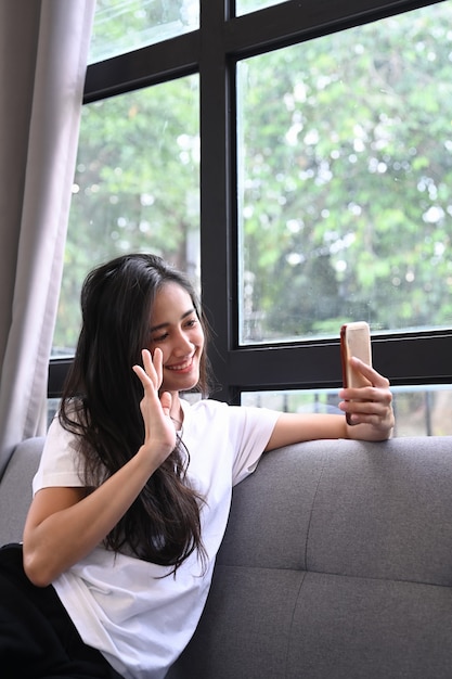 Retrato de dama segura que sostiene el teléfono inteligente haciendo selfies mientras está sentado en el sofá en casa.