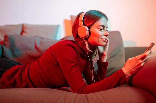 Retrato de una dama sonriente acostada en el sofá con auriculares y teléfono celular en las manos mientras escucha música en casa