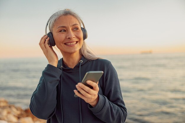 Retrato de una dama sonriente con capucha parada junto al mar con un teléfono inteligente y escuchando música