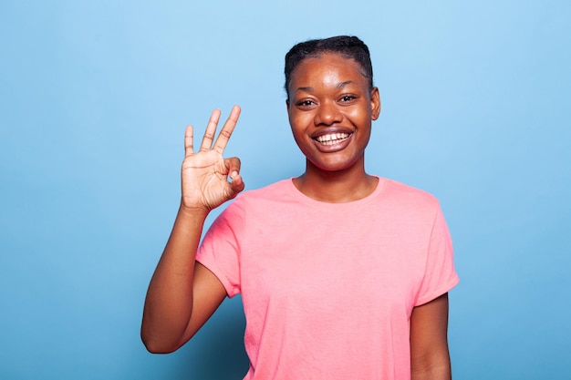 Retrato de adolescente afro-americano alegre positivo sorrindo para a câmera mostrando okey sinal de pé no estúdio com fundo azul. A jovem feliz otimista aprova a ideia. Sinal de acordo