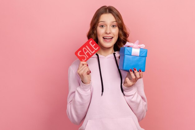 Retrato de adolescente alegre de cabelos cacheados com capuz segurando a palavra de venda e caixa de presente embrulhada preço baixo em presentes com desconto Foto de estúdio interior isolada em fundo rosa