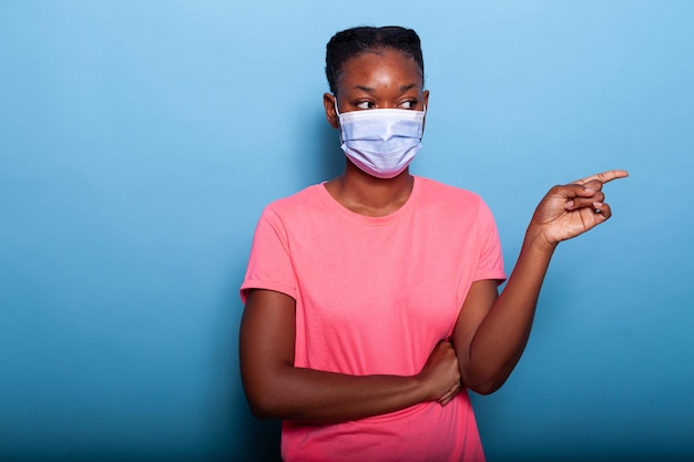 Retrato de adolescente americano africano com máscara facial protetora médica apontando o dedo para o lado direito em pé no estúdio com fundo azul. jovem posando para a câmera