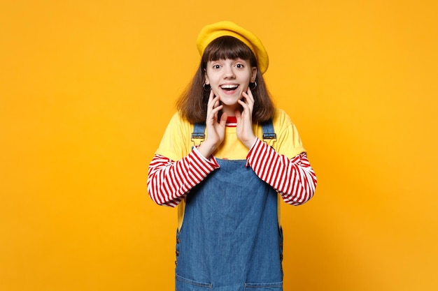 Retrato de adolescente animado em boina francesa e vestido jeans colocando as mãos no rosto isolado no fundo da parede amarela no estúdio. emoções sinceras de pessoas, conceito de estilo de vida. mock up espaço de cópia.