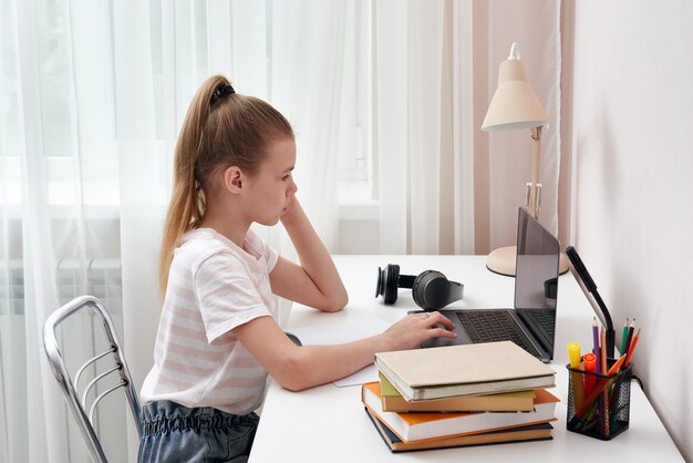 Retrato de adolescente aprendendo on-line com fones de ouvido e laptop tomando notas em um caderno, sentado em sua mesa em casa fazendo lição de casa