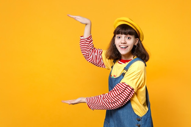 Retrato de adolescente excitada em boina francesa e vestido de denim apontando as mãos de lado isoladas no fundo da parede amarela no estúdio. Emoções sinceras de pessoas, conceito de estilo de vida. Mock up espaço de cópia.