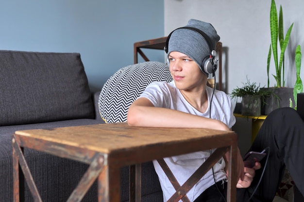 Retrato de adolescente hippie com chapéu com fones de ouvido, sentado no chão em casa. Cara bonito na moda olhando para o lado, copie o espaço. Juventude, estilo de vida e lazer