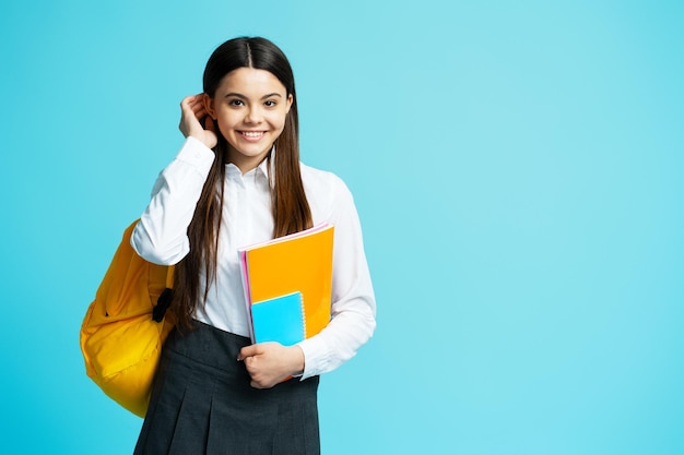 Retrato de adolescente positivo vestindo mochila amarela de uniforme escolar olhando para câmera fixando cabelo isolado em fundo azul de volta à escola