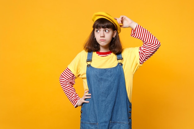 Retrato de adolescente preocupado na boina francesa, vestido jeans colocando a mão na cabeça, olhando de lado isolado em fundo amarelo. emoções sinceras de pessoas, conceito de estilo de vida. mock up espaço de cópia.