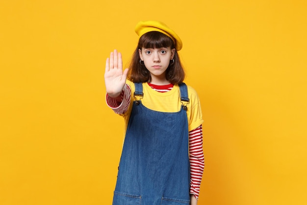 Retrato de adolescente séria na boina francesa, vestido jeans mostrando gesto de parada com palma isolada em fundo amarelo em estúdio. Conceito de estilo de vida de emoções sinceras de pessoas. Mock up espaço de cópia.