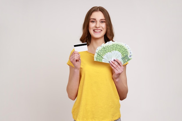 Retrato de adolescente sorridente com cabelo castanho em camiseta casual amarela segurando cartão de crédito e muito dinheiro sendo feliz por depósitos em dinheiro Tiro de estúdio interior isolado em fundo cinza