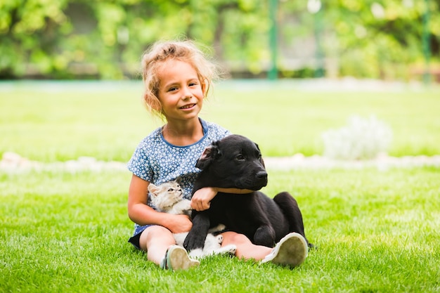 Retrato de adorável garotinha sorridente abraçando cachorro preto e gatinho branco melhores amigos da menina durante a quarentena e bloqueio passando tempo com animais de estimação ao ar livre no parque