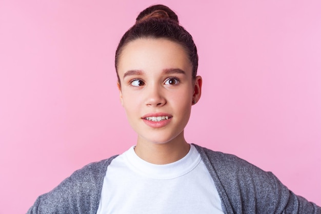 Retrato de adorável menina adolescente morena engraçada com penteado de coque em roupas casuais brincando com os olhos cruzados, se divertindo, rosto positivo infantil. tiro de estúdio interior isolado no fundo rosa