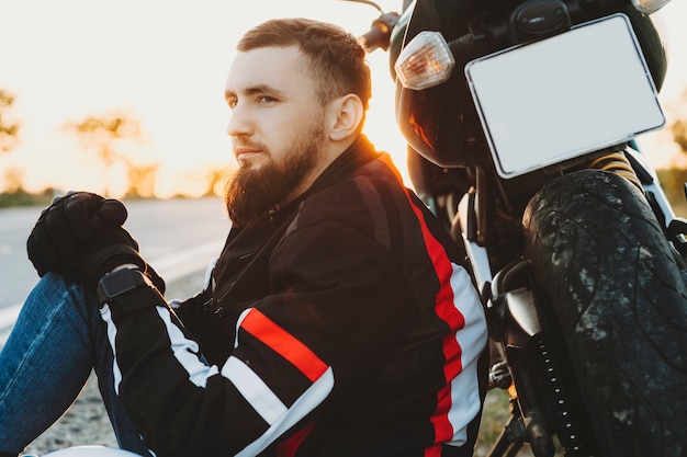 Retrato de adulto barbudo motociclista apoiado em sua bicicleta enquanto está sentado olhando para longe enquanto viaja de moto.