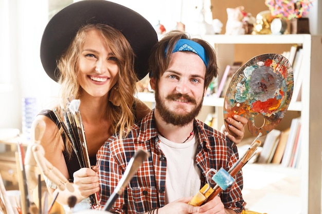 Retrato de alegre e feliz artista de cabelos longos femininos com chapéu bonito e artista masculino barbudo em bandana azul segurando pincel e paleta