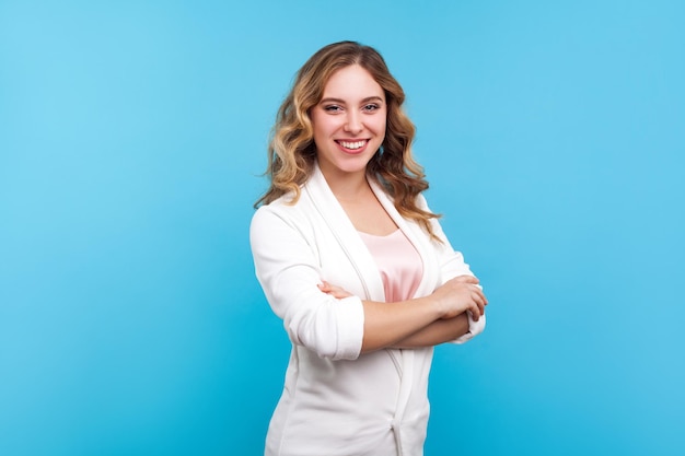 Retrato de alegre empresária brilhante com cabelos ondulados na jaqueta branca sorrindo para a câmera, parecendo elegante confiante e bem sucedido com as mãos cruzadas. tiro de estúdio interior isolado, fundo azul