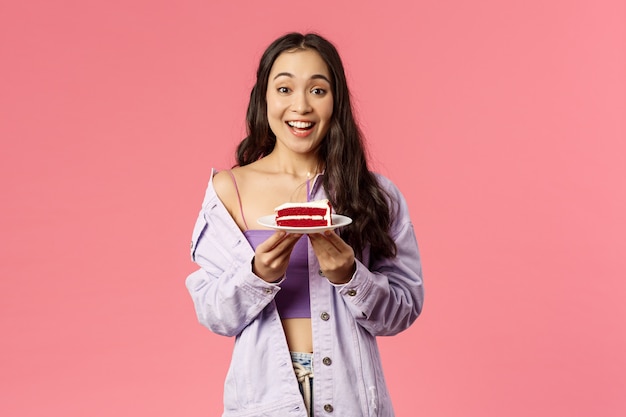 Retrato de alegre, feliz jovem bonito assou uma sobremesa especial para você, segurando o pedaço de bolo no prato, sugerindo experimentá-lo, sorrindo divertido e animado, comendo comida favorita, parede rosa